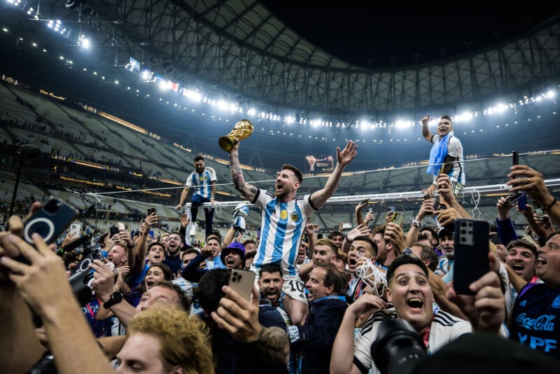 A soccer player in an Argentina jersey lifts the World Cup trophy, celebrating with jubilant teammates and fans inside a crowded stadium. Many people are cheering, raising hands, and taking photos with their phones.