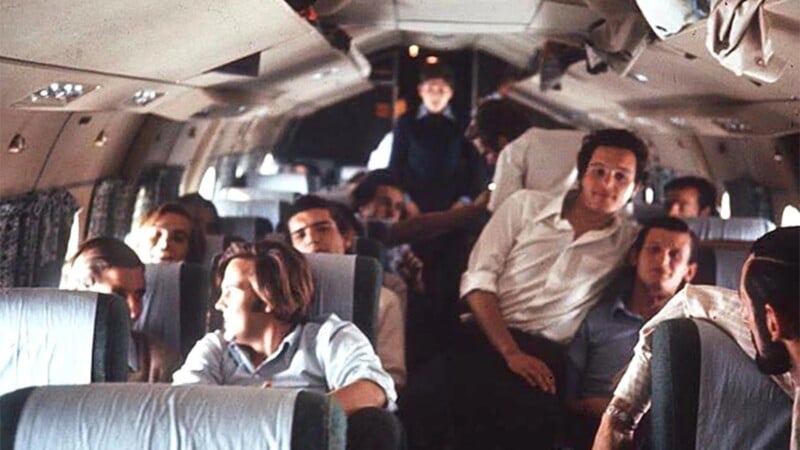 A group of young men sit and relax inside an airplane, some smiling and talking, with one person standing in the aisle towards the back. The atmosphere appears casual and friendly.