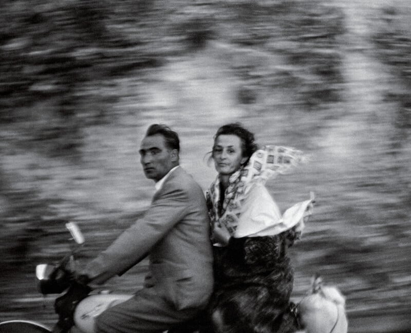 A black-and-white photo shows a man in a suit riding a motorcycle with a woman sitting behind him. The background is blurred, emphasizing their motion. The woman’s scarf and hair are flowing in the wind.