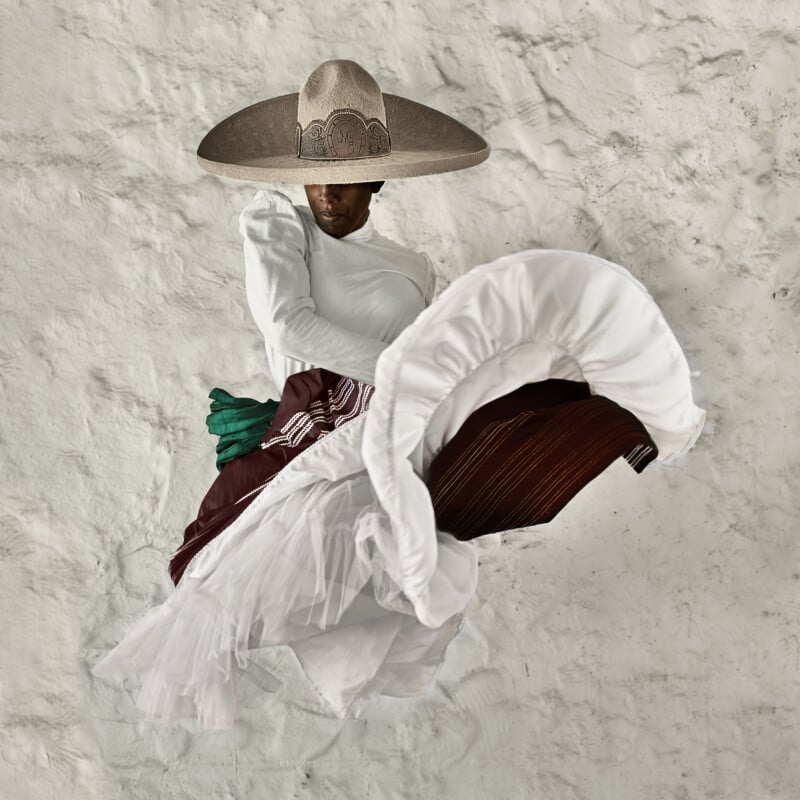 A dancer in a wide-brimmed hat and traditional folkloric attire leaps, lifting a white ruffled skirt, against a textured white wall.