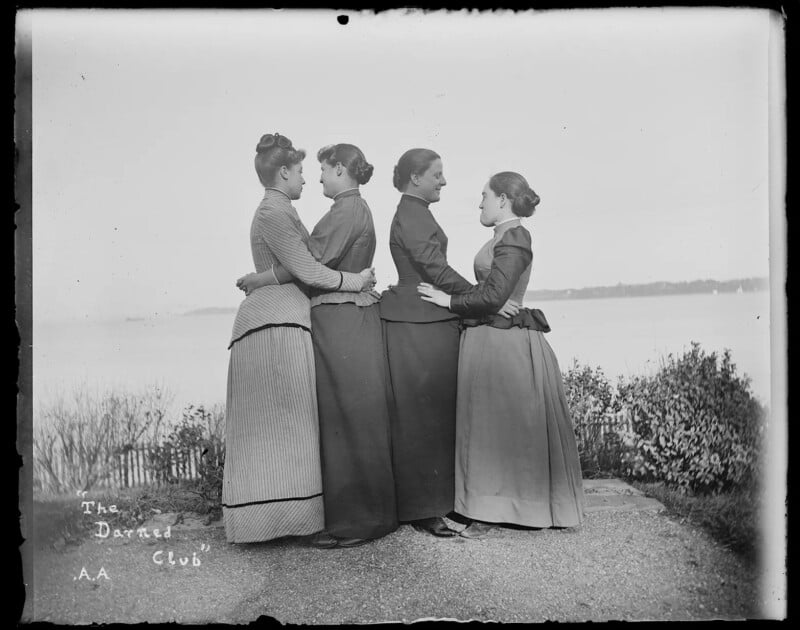 Four women in 19th-century dresses stand outdoors in a line, each facing and holding the woman in front of her. A body of water and sparse vegetation are visible in the background. The words “The Darned Club” are written on the photo.