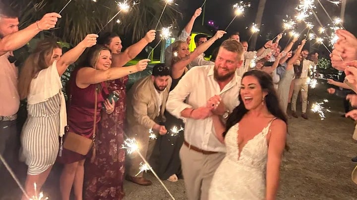 A joyful bride and groom walk hand-in-hand through a crowd of guests holding sparklers at night, celebrating their wedding. The guests smile and cheer as the couple passes by.