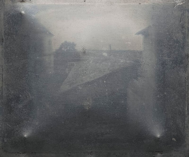 A faded, early black-and-white photograph shows a blurry rooftop scene with faint outlines of buildings and a tree in the background, framed by darkened, worn edges.