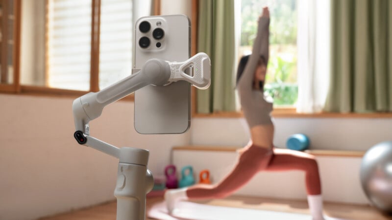 A smartphone mounted on a gimbal films a woman in workout clothes doing a yoga pose on a mat in a bright room with large windows, kettlebells, and an exercise ball.