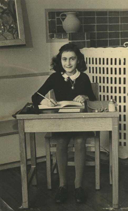 A young girl with dark hair sits at a wooden desk, writing in a notebook. She wears a dark dress with a white collar and smiles at the camera. The room has a radiator, a vase, and framed art in the background.