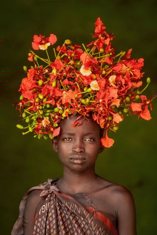 A young person with dark skin stands against a green background, wearing a large, elaborate headdress made of bright red and orange flowers with green buds. They have a calm, neutral expression and are draped in patterned fabric.