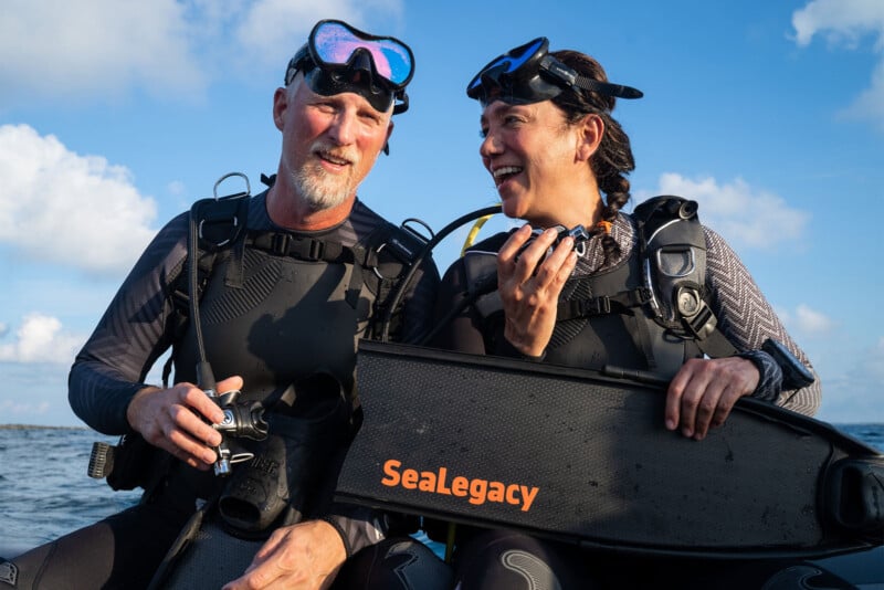 Two scuba divers in wetsuits and masks sit on a boat, smiling. One holds an underwater camera, the other holds a black fin labeled "SeaLegacy." The ocean and blue sky are visible in the background.