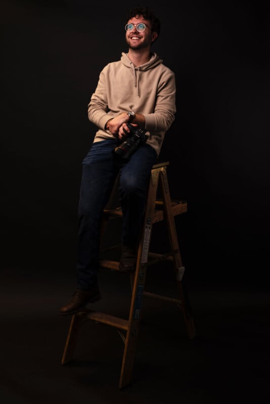 A man wearing glasses and a beige hoodie sits on a wooden ladder, holding a camera and smiling while looking upward, against a dark background.