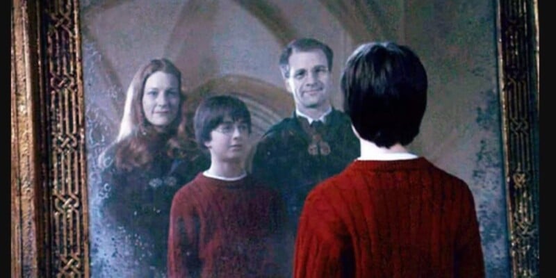 A boy in a red sweater stands in front of a large ornate mirror, seeing the reflection of himself with a smiling man and woman, who appear to be his parents, by his side.