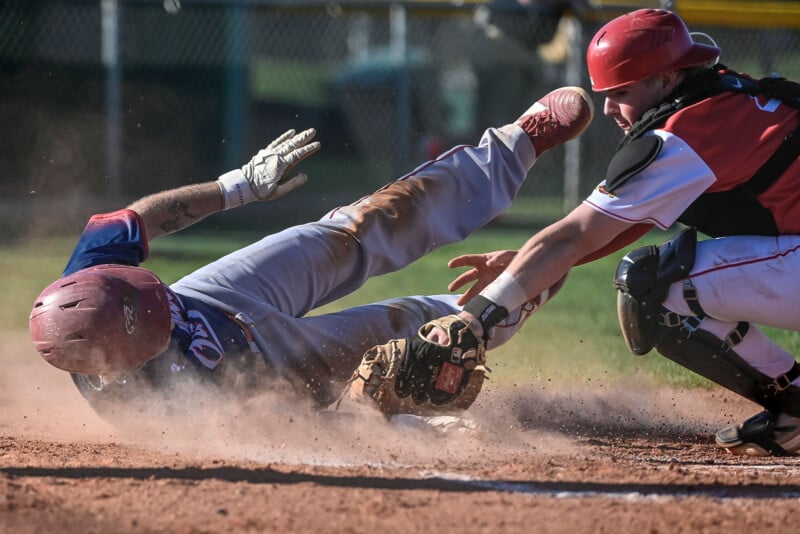 A baseball player slides into home plate as the catcher in red gear reaches to tag him, both surrounded by a cloud of dust on a sunny field.