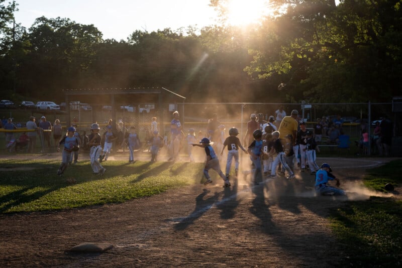 Children play baseball on a dusty field at sunset, with sunlight streaming through trees. Some kids run while others stand, and a cloud of dust rises near the base where a player slides in. Spectators watch from the background.