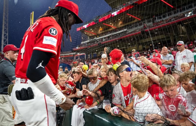 A baseball player in a red uniform signs autographs for excited young fans reaching over a barrier at a stadium, with many children holding balls, hats, and pens.