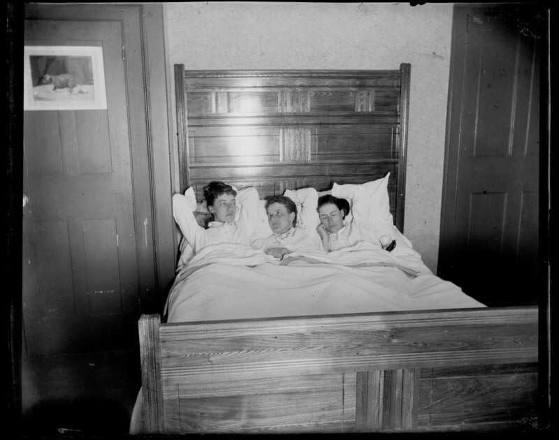 Three men in pajamas lie side by side in a large wooden bed, covered by a white sheet, in a dimly lit bedroom with two closed doors and a framed picture on the wall.