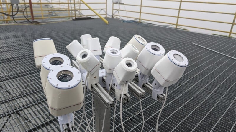 A group of white security cameras is mounted on metal brackets atop a grated platform, surrounded by yellow railings and industrial equipment. The cameras are angled in various directions and appear wet from rain.