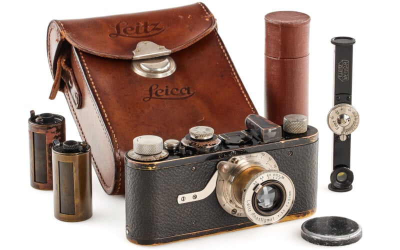 A vintage Leica camera with a leather carrying case, film canisters, a lens cap, a cylindrical container, and a rangefinder accessory displayed together on a white surface.