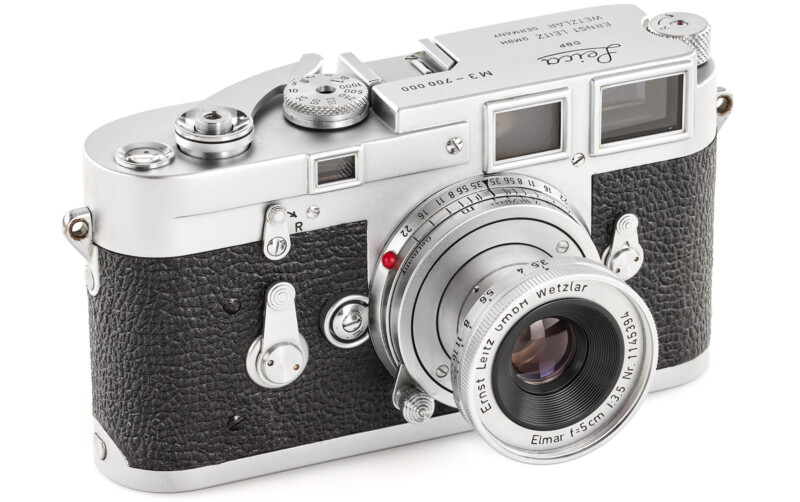 A vintage silver and black Leica film camera with a textured grip and an Ernst Leitz Wetzlar Elmar 50mm f/3.5 lens, resting on a white surface.