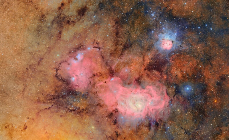 A vivid image of outer space shows colorful nebulae—pink, red, and blue clouds of gas and dust—surrounded by countless bright stars against a dark, speckled background.