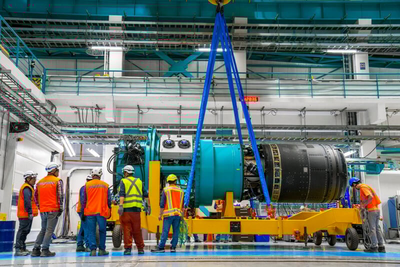 Workers in safety gear observe as a large jet engine is lifted by a crane in an industrial facility. The engine is attached to a yellow transport platform, with blue straps. The setting is bright, with pipes and equipment overhead.