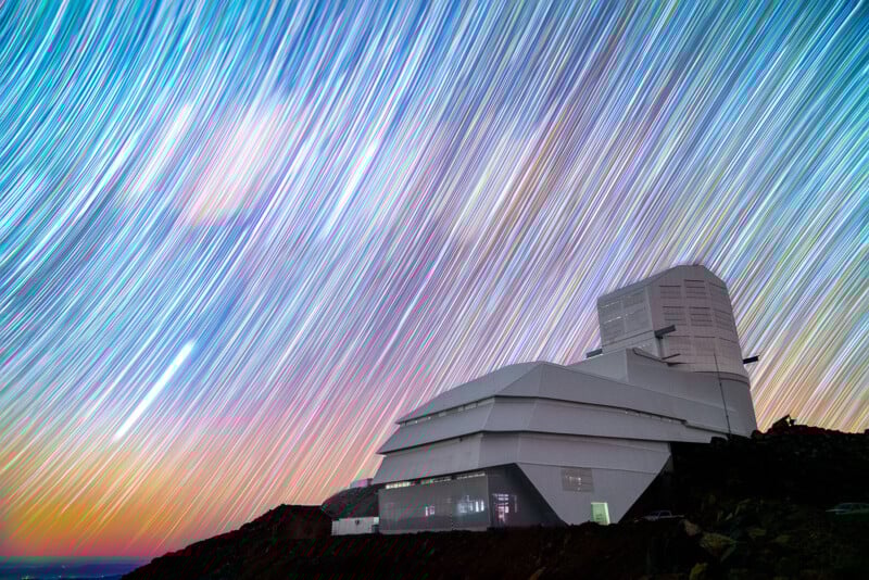 A modern observatory sits on a rocky hill under a night sky filled with colorful, streaking star trails, creating a vibrant, dynamic pattern above the building.