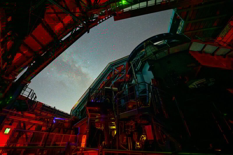 A night view from inside a large observatory, showing the telescope structure illuminated with red and green lights, with the star-filled sky and part of the Milky Way visible through the open roof.