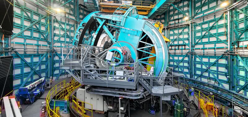 A large, blue observatory telescope surrounded by metal platforms and railings inside an industrial-looking facility with green steel framework and various equipment.
