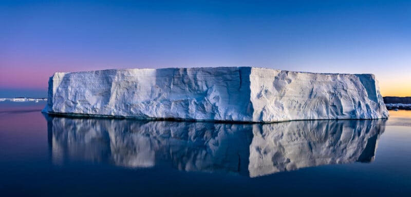 A large, flat-topped iceberg floats on calm, reflective water at sunset or sunrise, with a clear sky transitioning from deep blue to soft pink and yellow near the horizon.