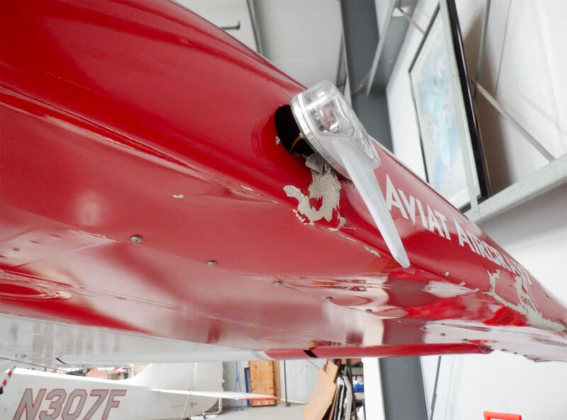 Close-up view of a damaged red aircraft wing with chipped and peeling paint near a navigation light. Part of another aircraft and a framed map are visible in the background inside a hangar.