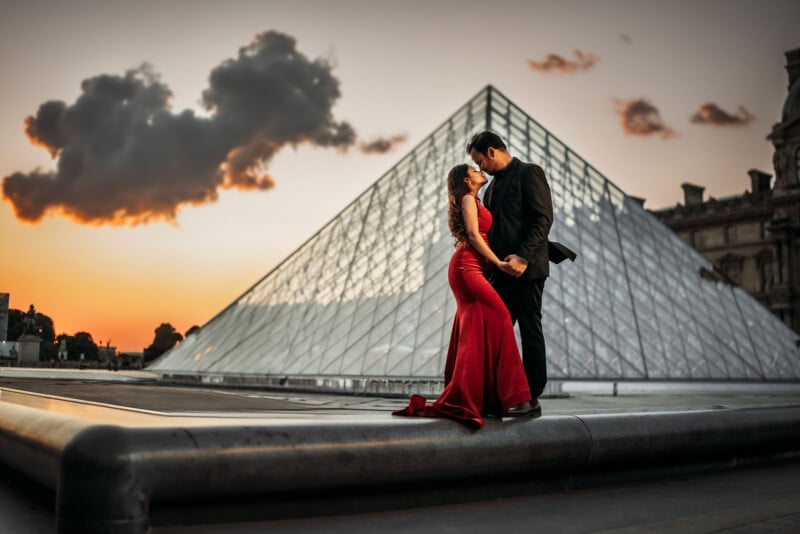 A couple dressed formally embrace and gaze at each other in front of the glass pyramid at the Louvre Museum in Paris at sunset, with a dramatic cloud and colorful sky in the background.