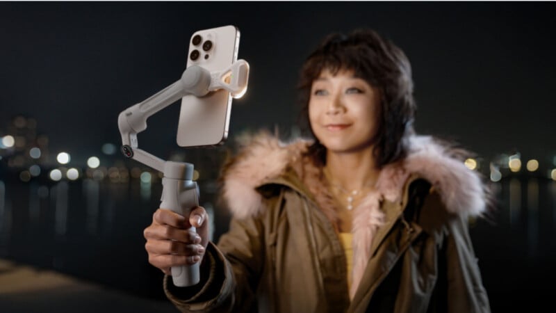 A person wearing a parka with a fur-lined hood holds a smartphone mounted on a gimbal stabilizer at night, with city lights blurred in the background.