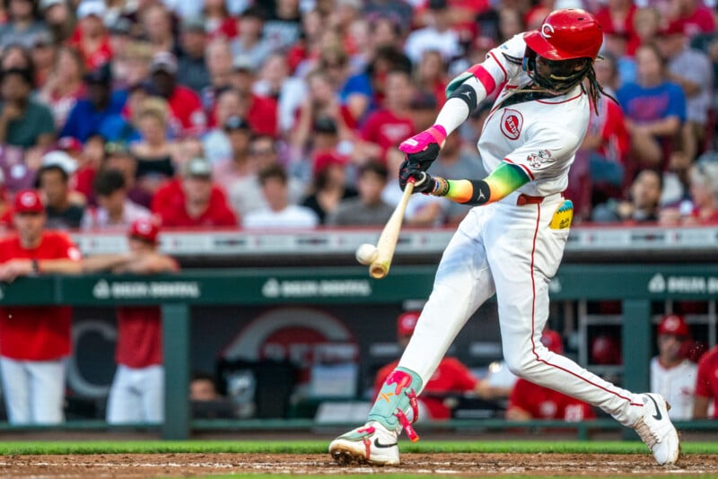 A baseball player in a white and red uniform swings a bat to hit a ball during a game, with a blurred crowd watching in the stadium seats in the background.