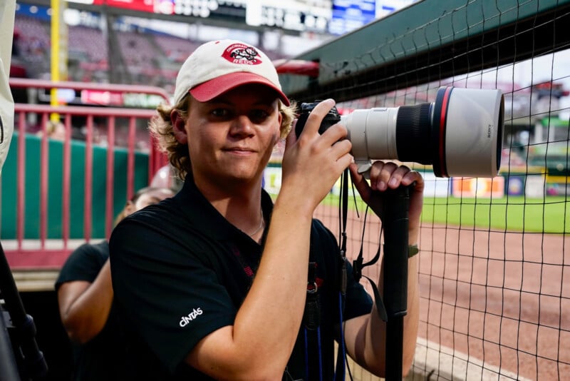 A young person wearing a baseball cap and black shirt holds a professional camera with a large lens at a baseball stadium, standing near a netted area by the field.