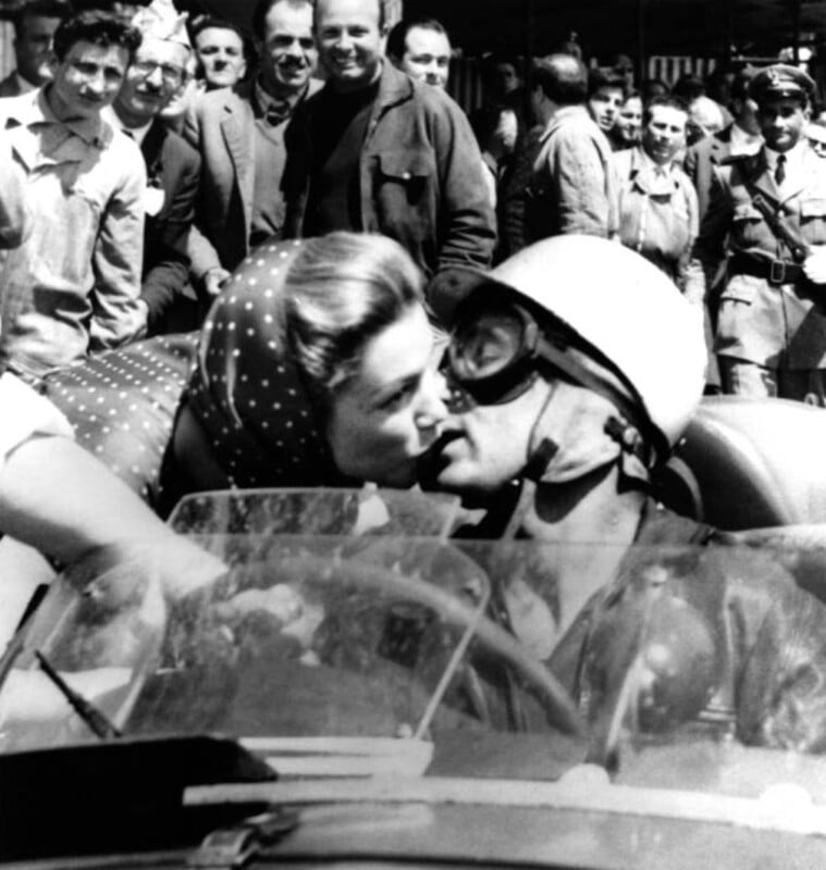 A woman wearing a polka-dot scarf kisses a man in a helmet and goggles sitting in a race car, while a crowd of people watches and smiles in the background.
