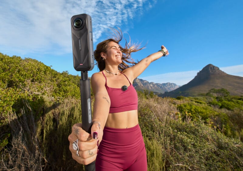 A woman in athletic wear smiles and holds a 360-degree camera on a selfie stick outdoors, with green bushes and mountains under a bright blue sky.