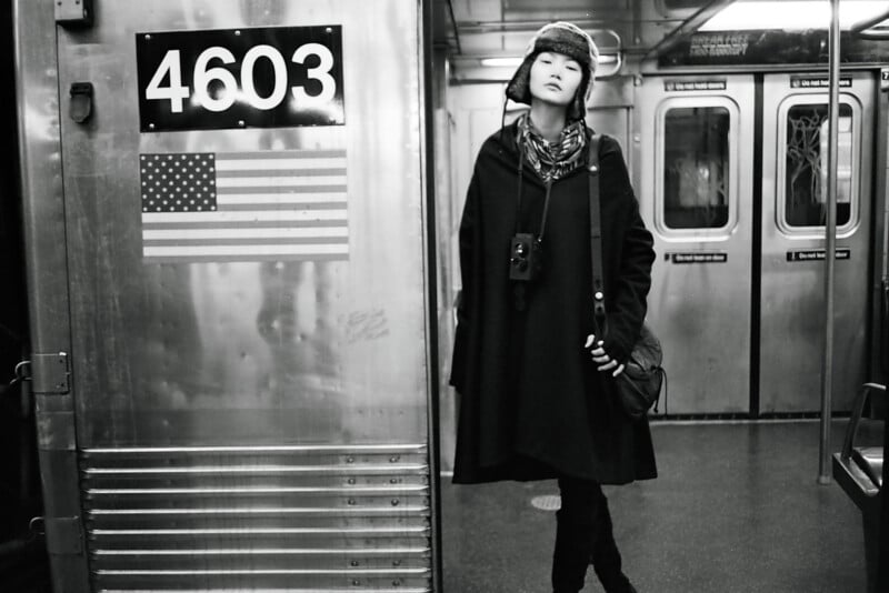 A person wearing a winter hat, scarf, and long coat stands leaning inside a subway car next to a metal wall labeled "4603" with an American flag. The subway interior is empty and lit with overhead lights.