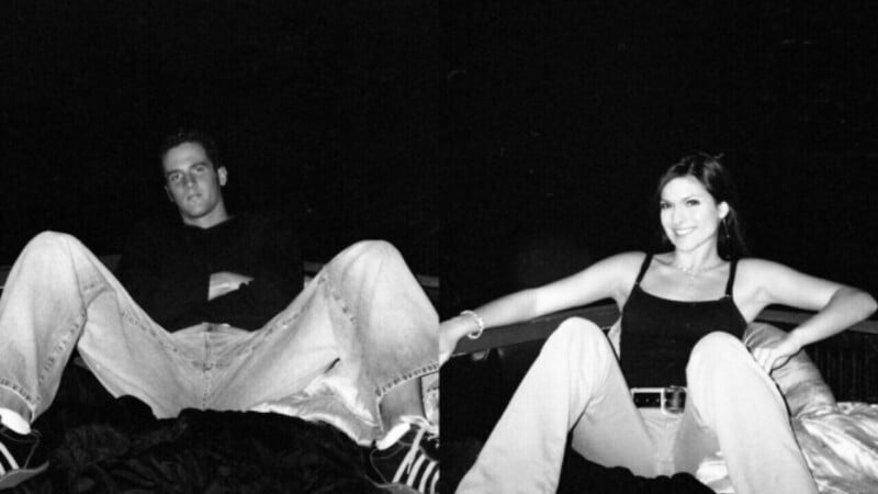 A black-and-white photo showing a man and a woman sitting separately with legs spread wide, arms crossed, and relaxed expressions. Both wear casual clothes and sit against a dark background.