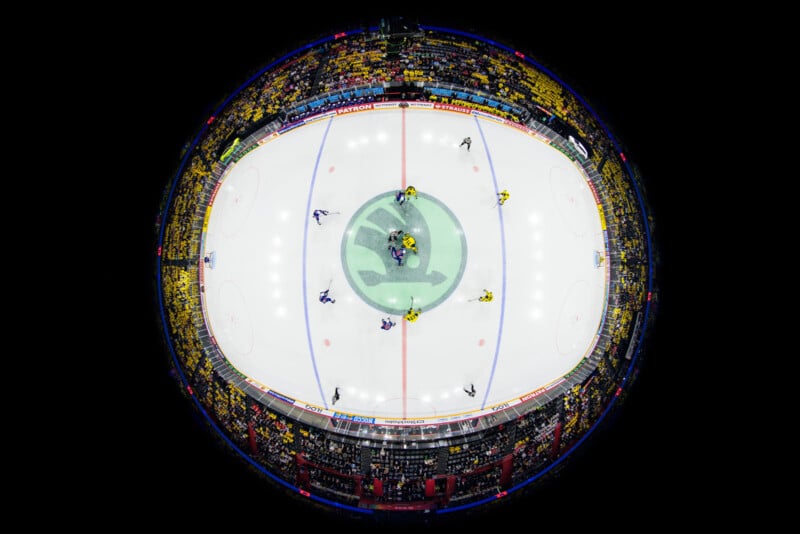 A fisheye lens view of an ice hockey arena packed with fans, showing players gathered at center ice for a face-off, with bright lights illuminating the rink and stands.