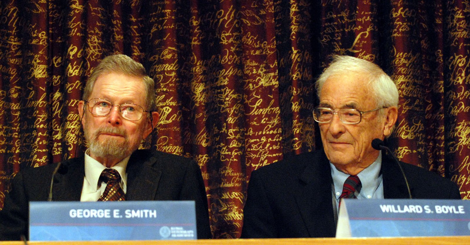 Two elderly men in suits sit at a table with microphones in front of a backdrop with gold cursive writing. Nameplates identify them as George E. Smith and Willard S. Boyle. Both appear attentive and composed.