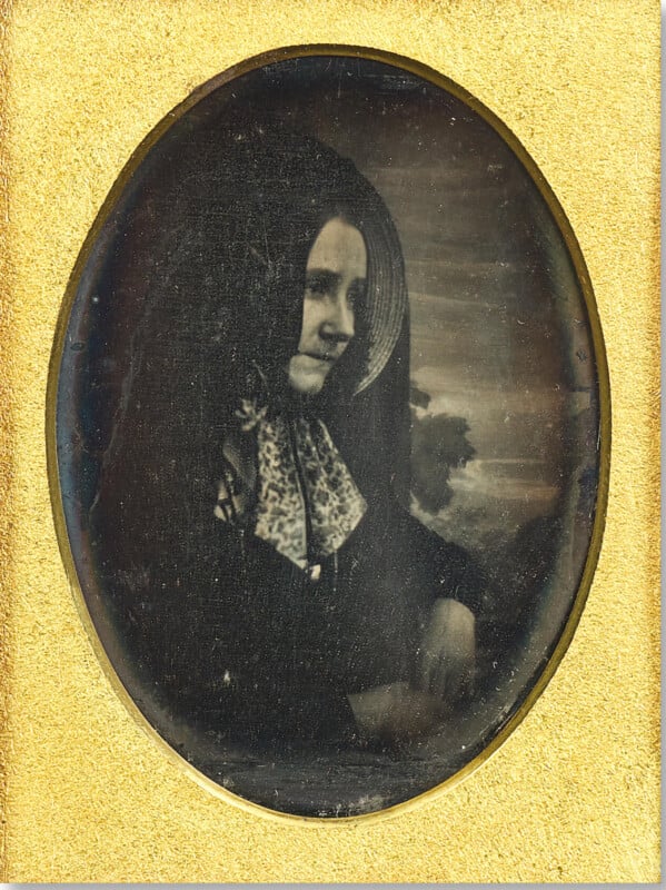A vintage black-and-white oval photograph of a woman in a dark dress with a scarf, framed by a gold mat. She is seated and looking slightly to the side with a neutral expression.