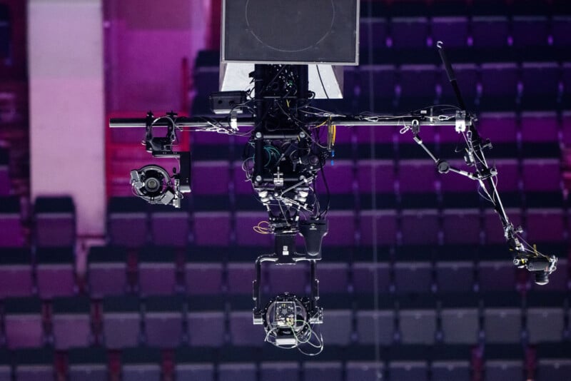 A close-up of a suspended camera rig with multiple cameras and cables, set against a blurred background of empty stadium seats.