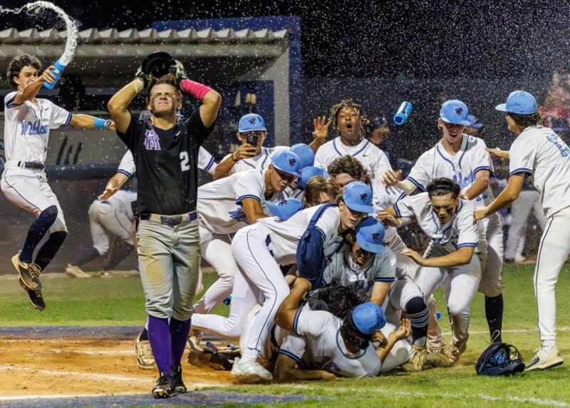 A group of baseball players in white uniforms celebrate joyfully on the field, piling on top of each other as one opposing player in a dark uniform stands nearby with his hands on his head looking disappointed.