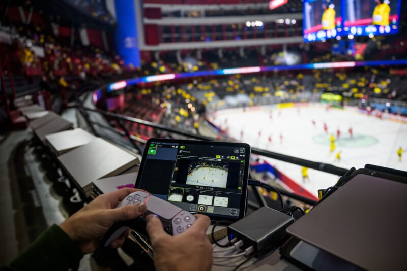 A person uses a PlayStation controller and tablet to operate a camera overlooking an ice hockey game in a large, brightly lit arena filled with spectators.