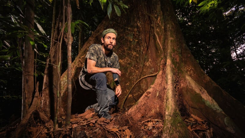 A man wearing camouflage clothing and a green headband crouches beside the massive roots of a large tree in a dense, leafy forest.