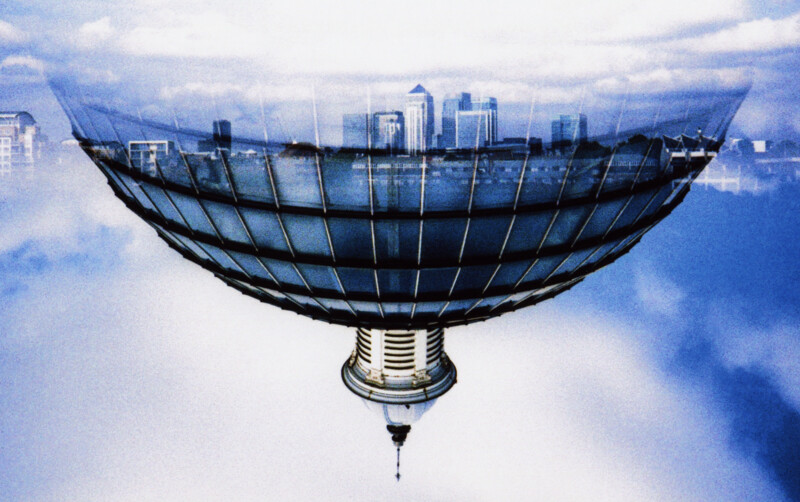 A surreal image of a cityscape reflected in a large, bowl-shaped glass structure, creating an optical illusion against a cloudy sky. The buildings and clouds blend together, forming a dreamlike, abstract scene.