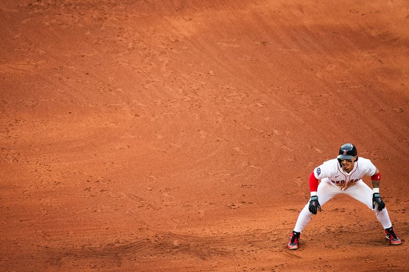 A baseball player in a white uniform and helmet stands ready in a defensive position on an orange-brown dirt field, with ample empty space around him.