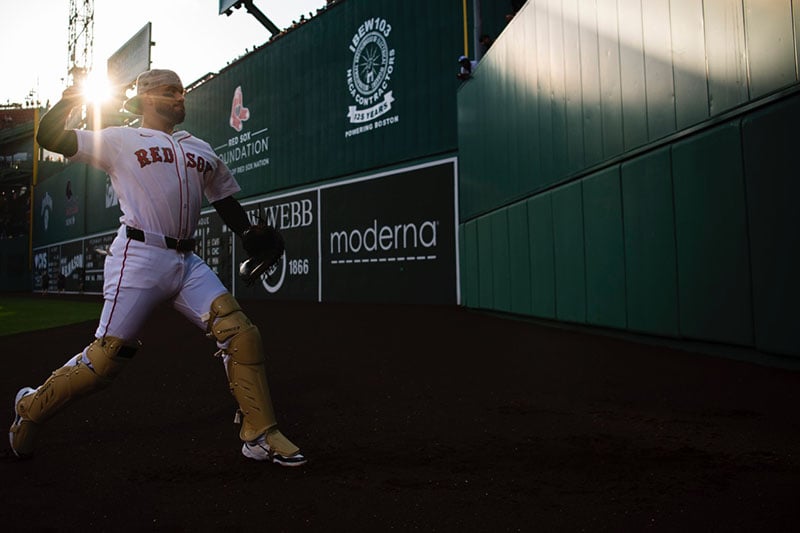 A baseball catcher in Red Sox gear throws a ball on a sunlit field near a stadium wall covered with advertisements and team logos.
