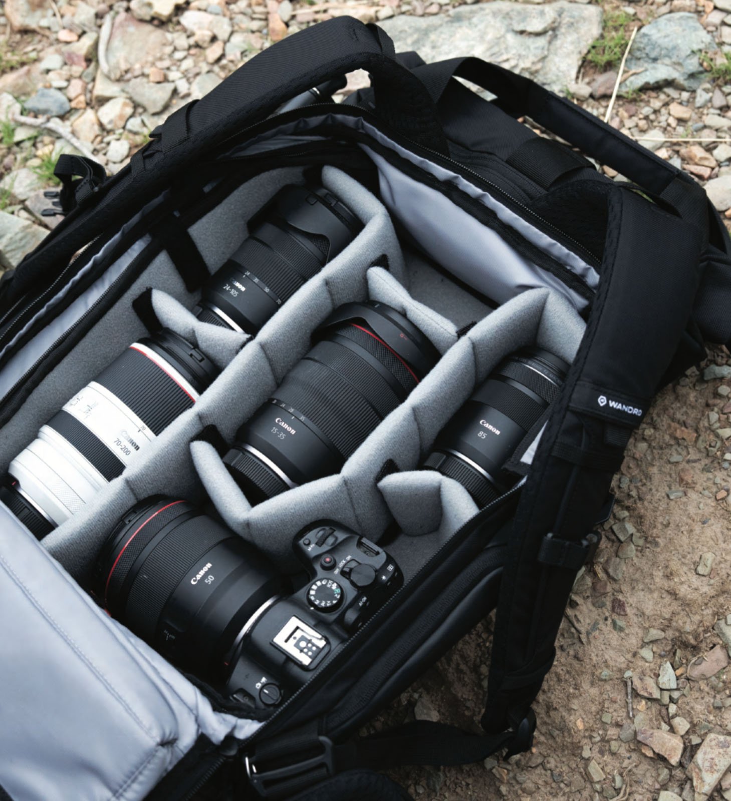A padded camera bag is open outdoors, showing a digital camera body and five interchangeable camera lenses neatly organized in separate compartments. The ground beneath has rocks and dirt.