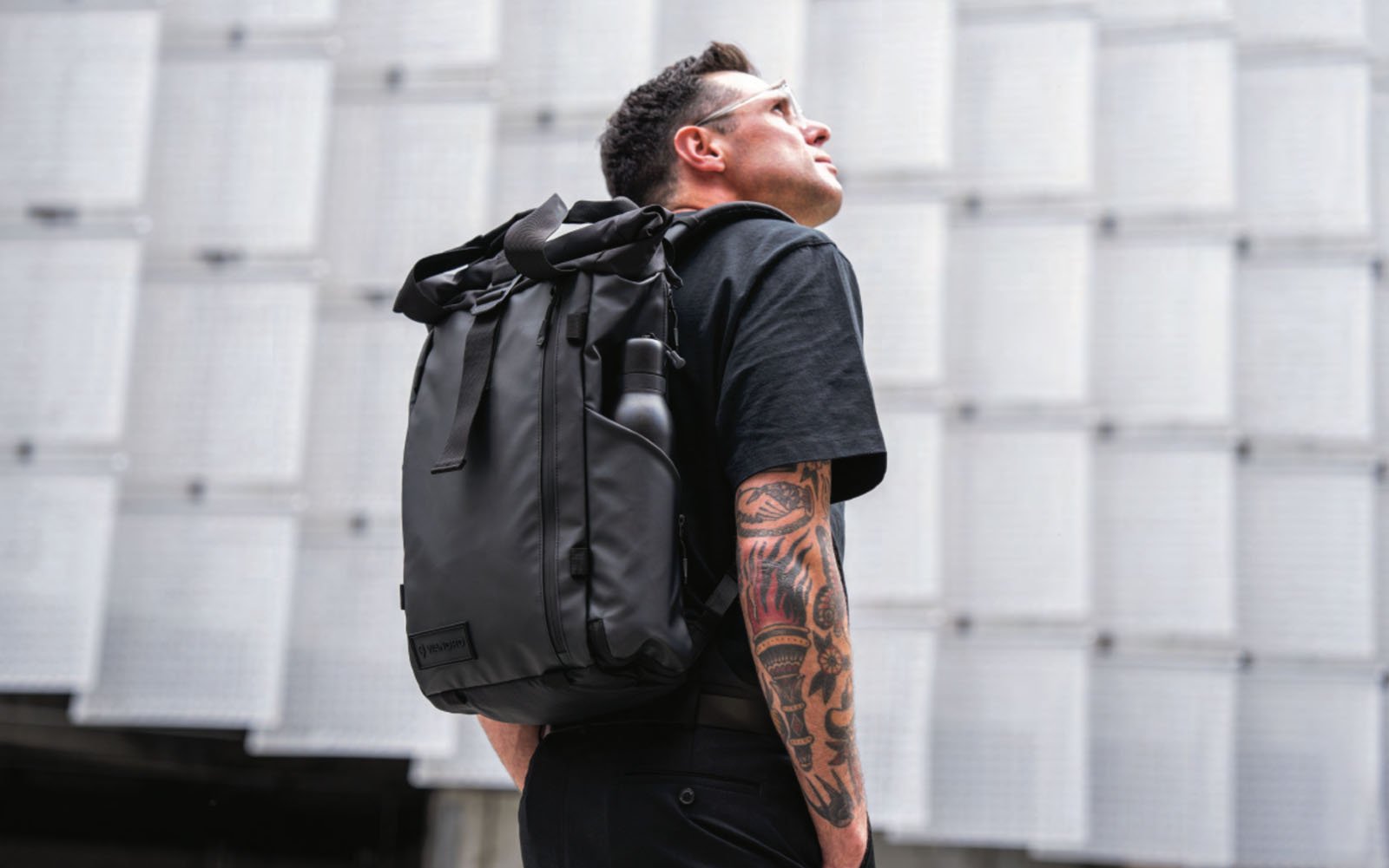 A man with tattooed arms, wearing sunglasses and a black t-shirt, looks up while carrying a large black backpack. He stands in front of a modern, geometric building facade.