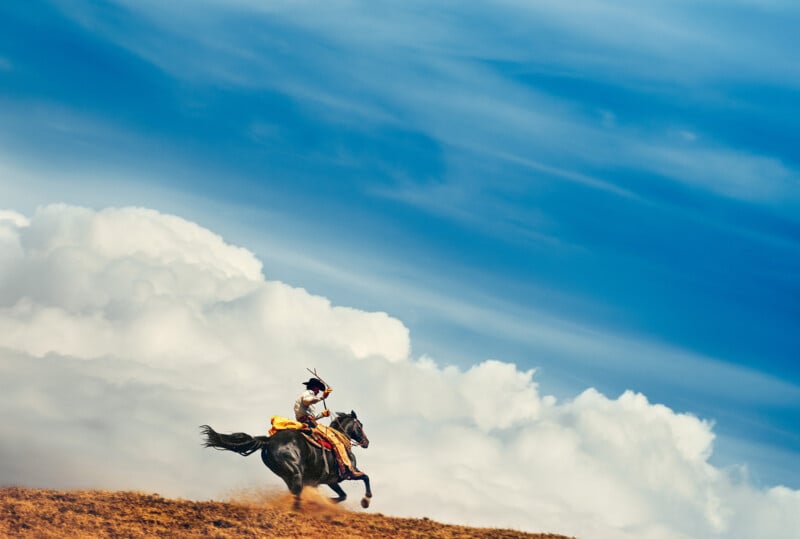 A person in traditional attire rides a galloping black horse across a dusty hill, with dramatic white clouds and a blue sky in the background.