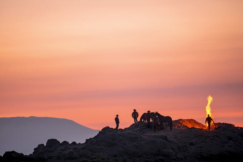 Silhouettes of people and horses stand on rocky terrain at sunset, with one person tending a campfire. The sky is painted in warm orange and pink hues, and distant mountains are visible in the background.