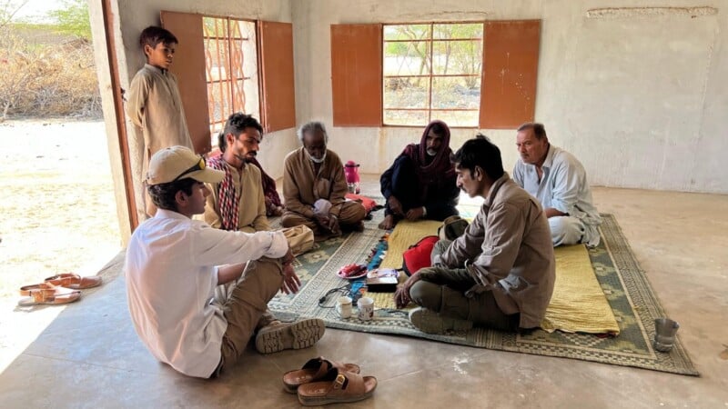 A group of seven people sit on a rug inside a simple, sunlit room with open windows, engaged in conversation. Some items, including cups and a book, are placed on the rug. Sandals are placed near the rug.